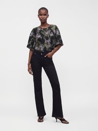 Femme portant un chemisier noir fleuri, un pantalon évasé noir et des sandales noires à talons, debout devant un fond blanc uni.