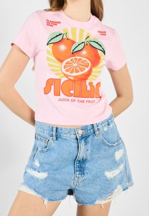 Osoba ubrana w różowy t-shirt z nadrukiem owoców cytrusowych i napisem "Sicilia Juice of the Fruit", w połączeniu z jasnoniebieskimi, podartymi szortami dżinsowymi.