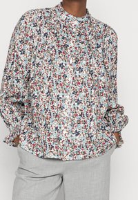 Blusa a maniche lunghe con motivo floreale, colletto a bande, maniche arricciate e bottoni, caratterizzata da colori come blu, rosso e beige su uno sfondo chiaro.
