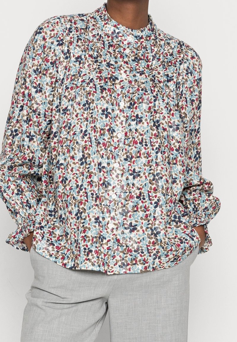 Blusa a maniche lunghe con motivo floreale, colletto a bande, maniche arricciate e bottoni, caratterizzata da colori come blu, rosso e beige su uno sfondo chiaro.