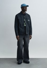 Μαύρο λειτουργικό μπουφάν, μαύρο γραφικό t-shirt, τζιν παντελόνι με φαρδιά μπατζάκια σε σκούρο χρώμα, παπούτσια με μαύρο μοτίβο και ανοιχτό μπλε τζιν καπέλο.