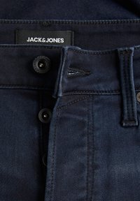 Jean en denim bleu foncé de Jack & Jones, présentant une fermeture à boutons sur le devant, une braguette zippée et des détails surpiqués autour des poches et des coutures.