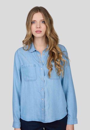 Chemise en denim bleu clair à manches longues, devant à boutons, deux poches poitrine et détails de surpiqûres visibles. Texture lisse et coupe décontractée.