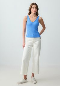 Canotta a coste azzurro chiaro con scollo a V, abbinata a pantaloni bianchi a gamba larga. Il modello indossa sneaker bianche. Outfit semplice e casual.