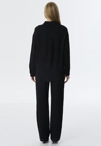Schwarzes, strukturiertes Longsleeve-Shirt mit Kragen, kombiniert mit weiten schwarzen Hosen und eleganten Absätzen. Einfaches Design, minimale Details.