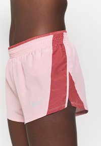 Primer plano de una persona que lleva pantalones cortos deportivos de Nike en color rosa y rojo, con paneles laterales perforados y un pequeño logo reflectante.