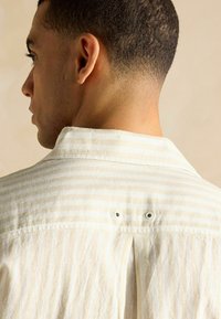 Küpsete varrukatega heledast kreemivärvi särgid, millel on vertikaalsed beežid triibud, plekitud seljaosa ja kaks nööbikohtadega kaelaosa.