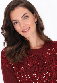 Donna con capelli castani ondulati, occhi blu e orecchini, che sorride, indossando un maglione rosso decorato con paillettes rosse e rosa.