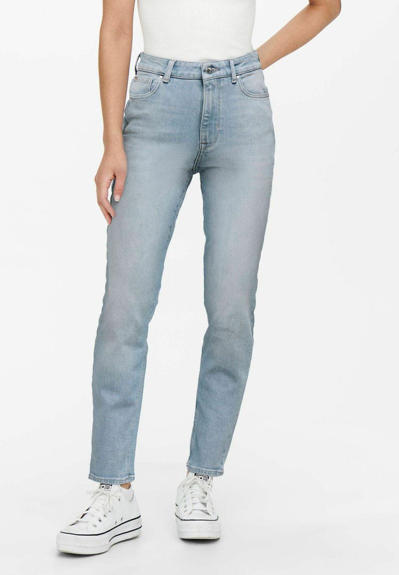 Personne portant un jean slim bleu clair, un haut blanc sans manches et des baskets blanches basses sur fond blanc.