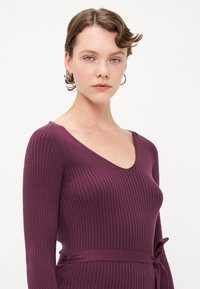 Lilla ribbestrikket kjole med lange ermer, v-hals og belte i midjen. Teksturen er myk, og passformen er tettsittende.