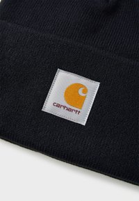 Black knit beanie featuring a white square patch with a yellow logo and red text. The texture is soft and tightly woven.