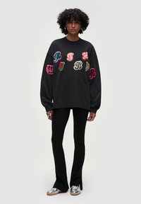 Felpa nera con lettere e motivi ricamati colorati sulle maniche, abbinata a pantaloni neri aderenti e scarpe argentate.