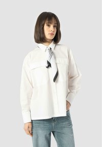 Weißes Hemd mit Knöpfen, das zwei Brusttaschen, lange Ärmel und einen schwarz-weißen gemusterten Schal am Hals hat. Blaue Jeans mit dezenten Details.