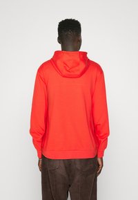 Felpa rossa con cappuccio, dal taglio rilassato, texture morbida e polsini a costine. Presenta una tasca a marsupio e un orlo piatto in vita.