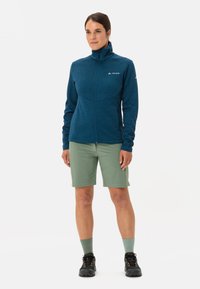 Giacca blu con cerniera, design testurizzato, maniche lunghe e tasche frontali, abbinata a pantaloni corti verdi e scarpe da trekking nere.
