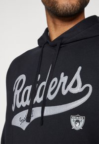 Juodas gobtuvinis megztinis pagamintas iš minkštos medžiagos, turi storą pilką siuvinėjimą su žodžiu „Raiders“ ir mažą komandos logotipo pleistrą priekyje.