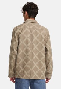 Beige jacket with a geometric pattern in a lighter shade. Features a collared neckline and a relaxed fit with button closures.