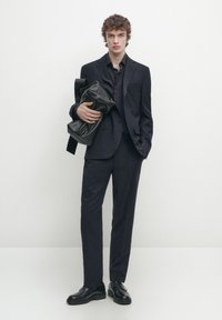 Tailleur scuro realizzato in tessuto liscio, accompagnato da una borsa nera strutturata. Presenta una silhouette aderente e scarpe nere lucide.