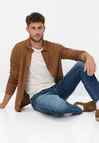 Cardigan marrone con bottoni sopra una t-shirt bianca, abbinato a jeans blu e scarpe marroni. Il modello è seduto con un braccio appoggiato sulla gamba.