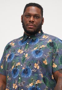 Camisa de botones de manga corta en tela azul oscura con vibrantes patrones de flores y hojas en verde, naranja y azul. Textura ligera.