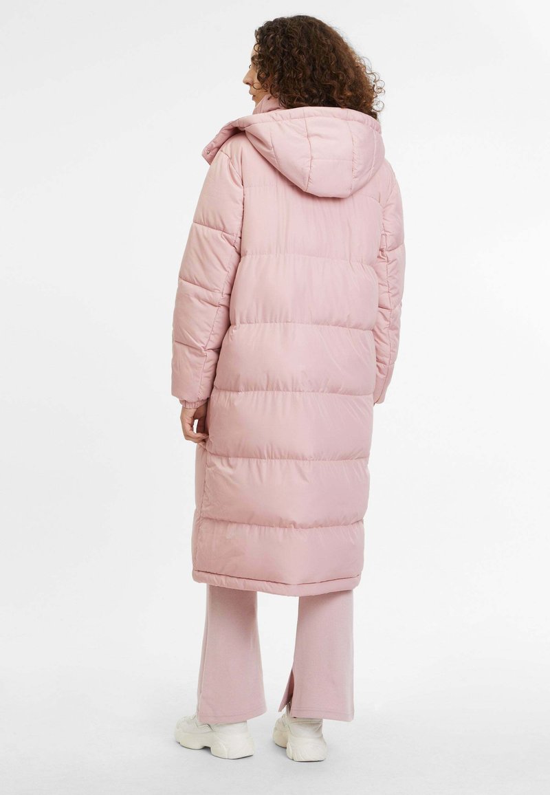 Lang roze gewatteerde jas met capuchon, met gestikte panelen en een relaxte pasvorm, gecombineerd met bijpassende broeken en lichtgekleurde schoenen.