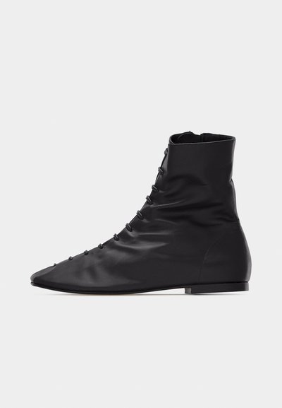 Bottine en cuir noir souple avec lacets latéraux, embout pointu, semelle plate et fermeture éclair à l'arrière.