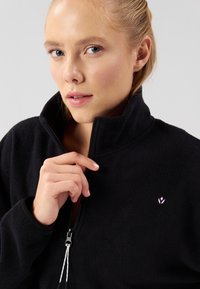 Černá fleecová bunda s vysokým límcem, polovičním zipem a malým růžovým logem na levé straně hrudi. Měkká textura a ležérní design.