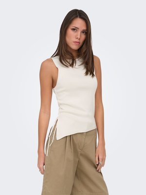 Femme aux cheveux bruns portant un haut blanc sans manches et un pantalon beige ample, debout devant un fond clair uni.