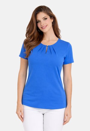 Kobieta o długich brązowych włosach, ubrana w niebieską bluzkę z krótkim rękawem, plisami i detalem z wycięciem, w połączeniu z białymi spodniami, uśmiecha się do kamery.