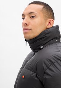 Schwarze Steppjacke mit hohem Kragen, silbernen Druckknöpfen und kleinem mehrfarbigem Logo auf der Brust. Glatte Textur.