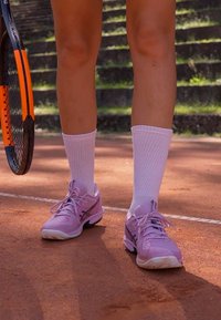 Világos rózsaszín sportcipők texturált anyaggal, szürke részletekkel és gumitalppal, fehér térd fölé érő zoknikkal párosítva, egy agyagpályán.