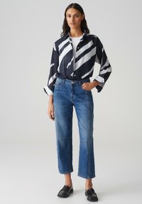 Camicia a righe bianche e nere con bottoni, jeans blu a vita alta e gamba dritta, e scarpe nere senza lacci, su uno sfondo neutro.