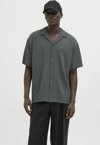 Camisa de manga corta, con botones, en tejido texturizado de color gris oscuro. Presenta un collar clásico y un ajuste holgado, combinada con pantalones negros y una gorra.