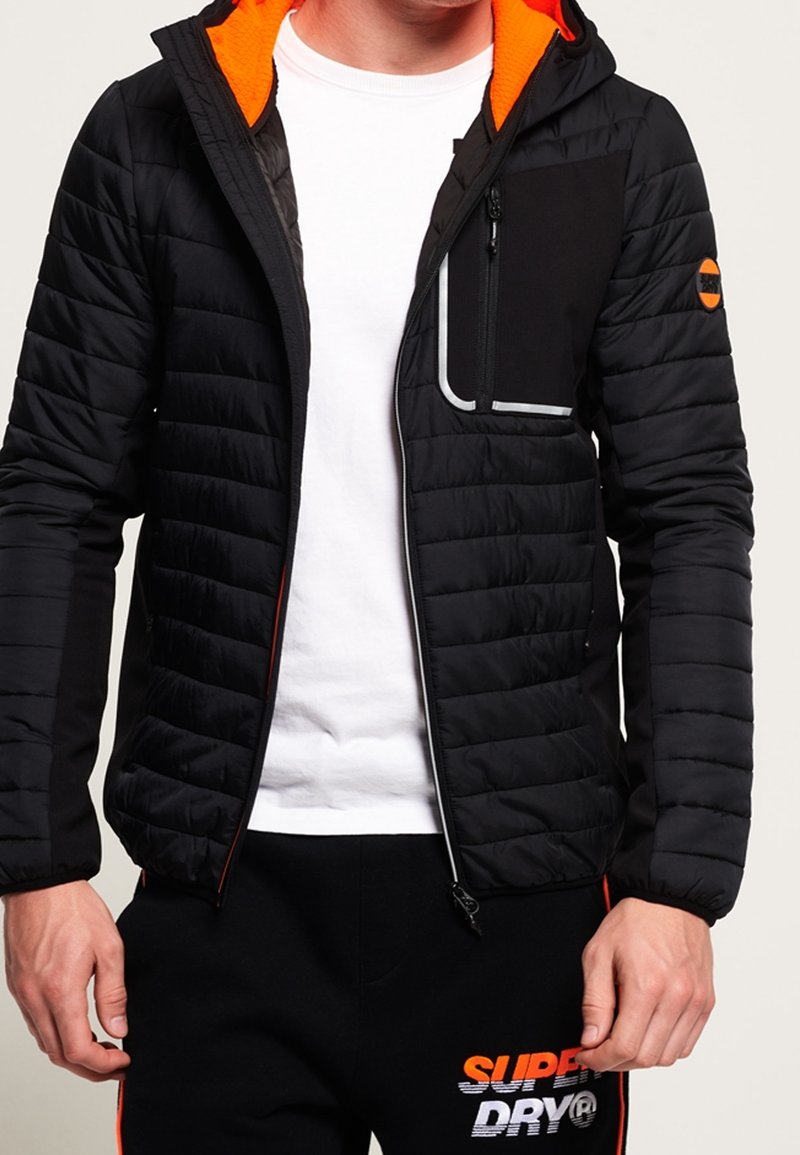 Schwarze gesteppte Jacke mit orangefarbenem Kapuzenfutter, schmaler Passform, gesteppter Textur, Frontreißverschluss, Seitentasche und schwarzem Akzentpaneel mit weißem Besatz.