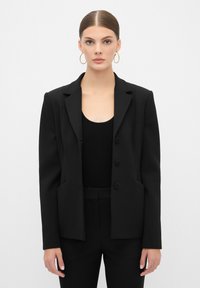 Svart skräddarsydd blazer med en strukturerad design, med spetsiga slag, tre knappar och sidfickor, bärs över en svart ribbad topp.