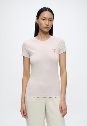 Femme aux longs cheveux noirs porte un t-shirt ajusté rose clair avec un logo Guess rouge sur la poitrine et un pantalon couleur crème.