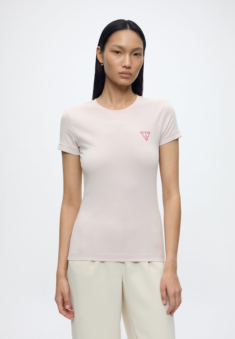 Femme aux longs cheveux noirs porte un t-shirt ajusté rose clair avec un logo Guess rouge sur la poitrine et un pantalon couleur crème.