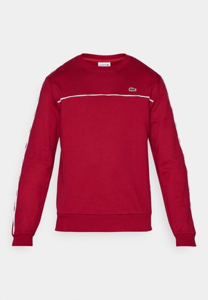 Červená mikina s dlouhými rukávy, vyrobená z bavlny. Obsahuje bílý horizontální pruh a malé zelené logo na hrudi. Nastavitelné manžety.