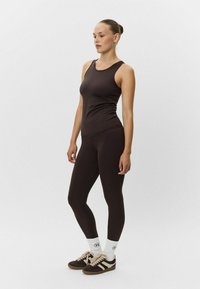 Hnedý bezrukávový top skombinovaný s hnedými legínami, oba vyrobené z hladkej tkaniny. Modelka má na sebe biele ponožky a hnedé tenisky so pruhmi.