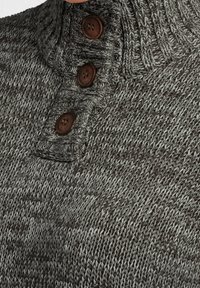 Pull en tricot avec un col montant, dans des tons gris présentant un motif texturé. Trois boutons marron au niveau du col ajoutent du détail.