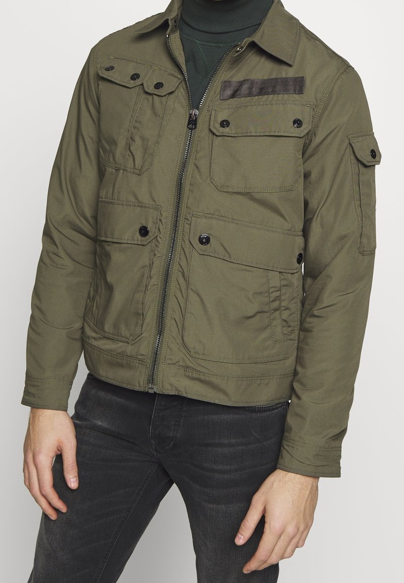 Olivgrön jacka med dragkedja, fyra framfickor och en texturerad brun axelpanel. Tillverkad av ett slätt tyg och har en krage.