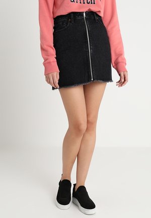 Person iført en sort denim mini nederdel med lynlås foran, lyserød langærmet top og sorte slip-on platform sneakers.