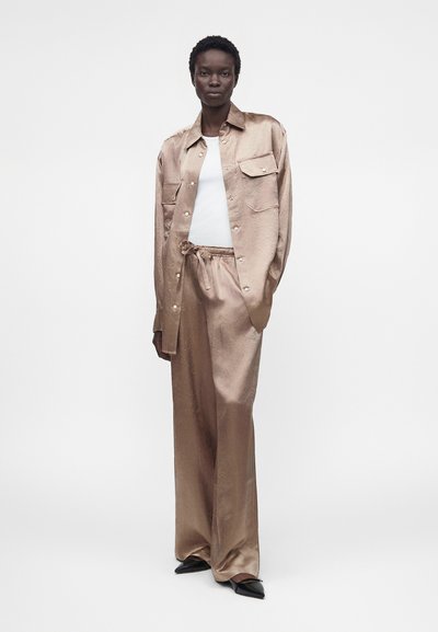 Το μοντέλο φοράει μια γυαλιστερή μπεζ σατέν πουκάμισο και παντελόνια με φαρδιά μπατζάκια, σε συνδυασμό με λευκό T-shirt και μαύρα μυτερά παπούτσια.