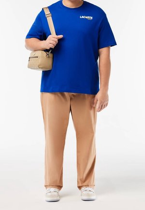 Kungsblå t-shirt med Lacoste-logotyp, beige byxor, beige axelväska och vita sneakers med ljusblå detaljer. Enfärgad bakgrund.