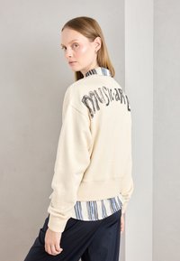 Sudadera crema con gráfico "musical" en la espalda, superpuesta a una camisa de cuello con rayas azules y blancas, combinada con pantalones oscuros.