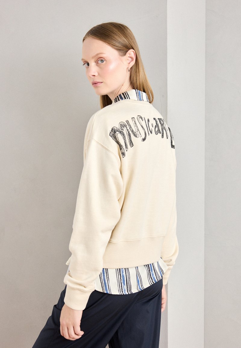 Sudadera crema con gráfico "musical" en la espalda, superpuesta a una camisa de cuello con rayas azules y blancas, combinada con pantalones oscuros.