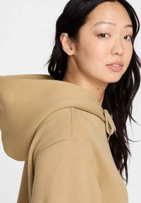 Haut à capuche beige avec un cordon de serrage, présentant une texture douce et une poche kangourou. Le design inclut des coutures visibles le long des joints.