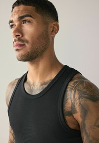 Black ribbed tank top with a round neckline, worn by a man showing defined tattoos on his chest and arm. Neutral background.