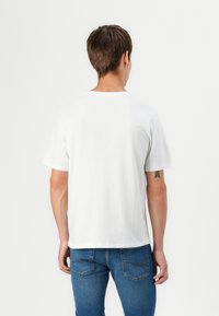 Hvit bomulls t-skjorte med korte ermer og rund hals, vist fra bak. Brukt med blå denim jeans med baklommer.