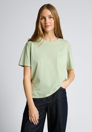 Vrouw met lang lichtbruin haar, gekleed in een lichtgroen t-shirt met zakken en donkerblauwe jeans, staand met een hand in haar zak.